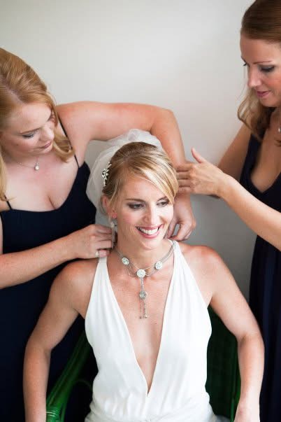Smiling bride in her wedding dress being helped with her veil and jewellery on the morning of her wedding