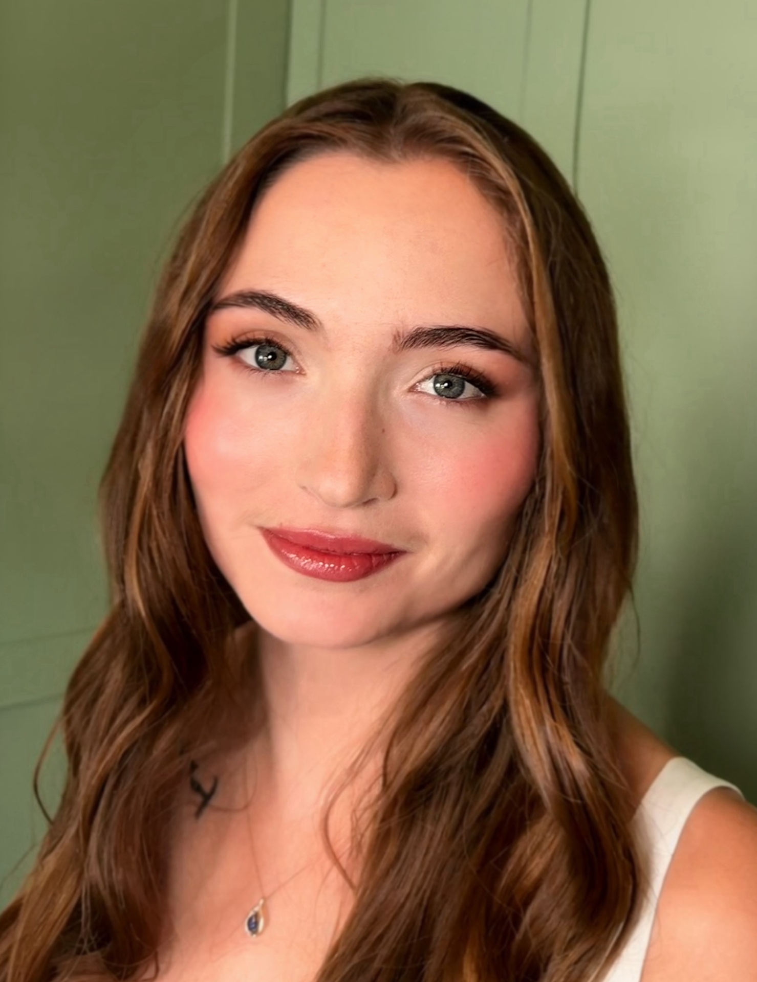 Soft glam makeup look with glowing skin, rosy cheeks and glossy lips against a sage-green panelled wall